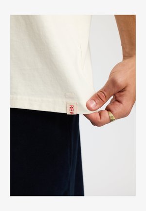 Lichtbeige katoenen t-shirt met een rechte zoom. Een klein rood label dat aan de zijkant is gestikt toont het woord "REV." De structuur lijkt glad.