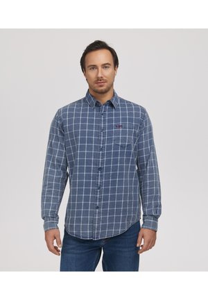 Mężczyzna w niebieskiej koszuli w kratę z długim rękawem na guziki i niebieskich dżinsach, stojący na jednolitym jasnym tle.