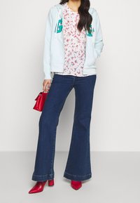 Sudadera azul claro con letras verdes, camisa blanca con flores, jeans acampanados oscuros y tacones rojos puntiagudos, sosteniendo un pequeño bolso rojo.