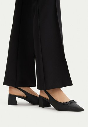 Pies con zapatos negros de tacón grueso y tira trasera con detalle de lazo, combinados con pantalones negros de pierna ancha sobre un fondo blanco.