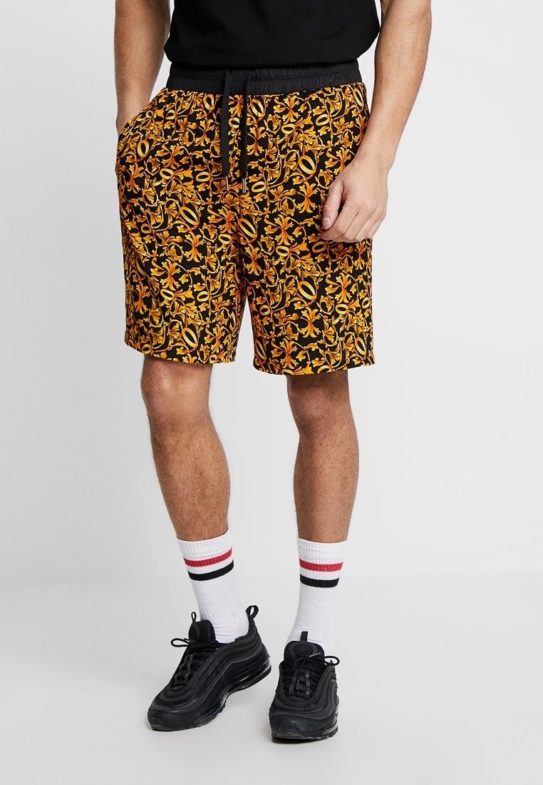 Homme portant un short à motif floral noir, orange et jaune, des chaussettes blanches avec des rayures rouges et noires et des baskets noires, debout les mains dans les poches.