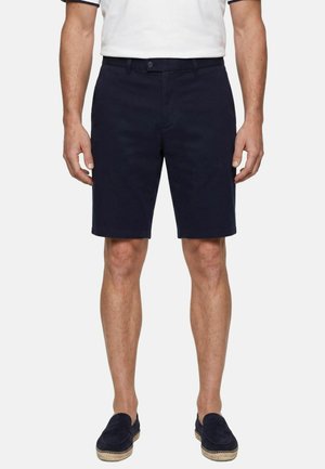 Man draagt donkernavy knielange shorts, witte korte mouwen shirt en donkere instapschoenen, staand tegen een witte achtergrond.