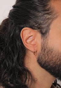 Pendiente de aro dorado en la oreja izquierda, sobre cabello largo, oscuro y ondulado, junto con una barba, mostrando la textura de la piel y el color natural del cabello.