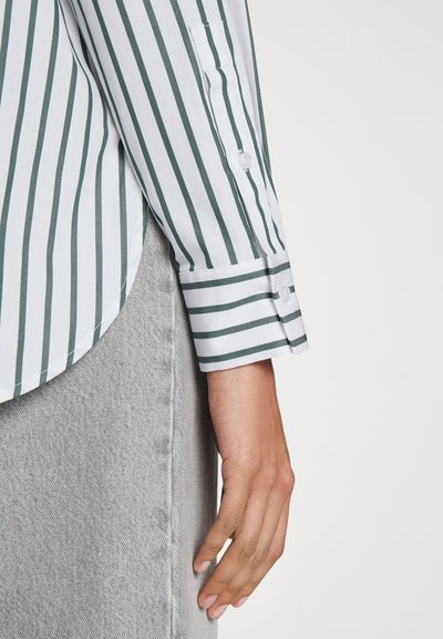 Camicia bianca con strisce verticali verde scuro; presenta un orlo arrotondato e polsini con bottoni, abbinata a una gonna in denim grigia chiara.