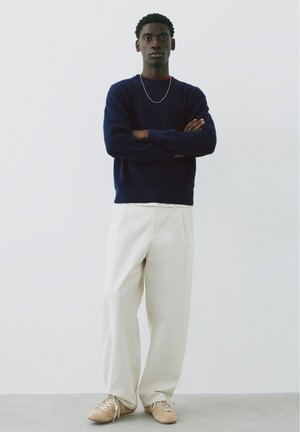 Jeune homme debout, bras croisés, portant un pull bleu marine, un pantalon blanc à jambes larges, des baskets beiges et un fin collier argenté sur un fond uni.