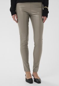 Pantalon ajusté, taille haute, en similicuir taupe lisse. Présente un design épuré et des jambes effilées, associé à des mules noires.