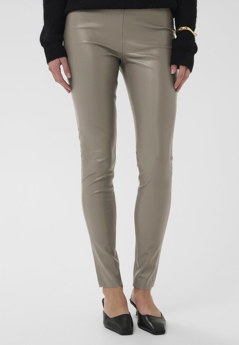 Pantalon ajusté, taille haute, en similicuir taupe lisse. Présente un design épuré et des jambes effilées, associé à des mules noires.