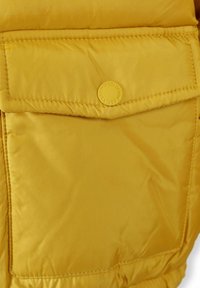 Section d'une veste matelassée jaune, présentant une grande poche avec un logo circulaire. Le matériau semble lisse avec un léger éclat.
