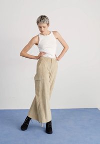 Beige cargo pants with wide legs, drawstring waist, and multiple pockets, paired with a white tank top and black lace-up boots.