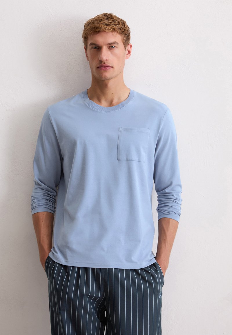 Camisa de manga larga de color azul claro con cuello redondo y bolsillo en el pecho, combinada con pantalones de rayas oscuros, de pie contra un fondo neutro.