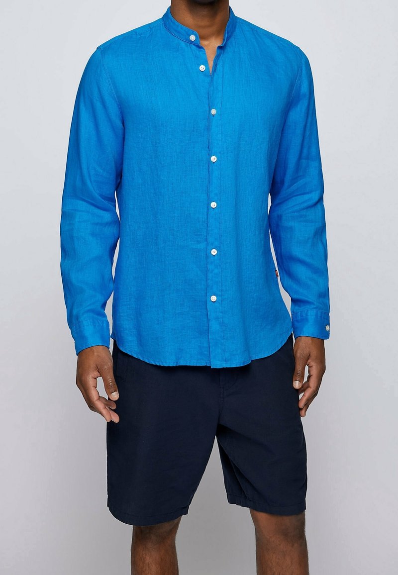 Homme portant une chemise bleu vif à manches longues avec col mandarin et un short bleu marine, debout devant un fond uni.