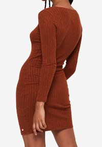 Femme portant une robe en tricot côtelé ajustée à manches longues de couleur rouille, se tenant de profil avec une main détendue à ses côtés.
