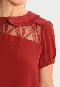 Blouse rouge avec un col à volants, des manches courtes bouffantes et un panneau en dentelle orné de motifs floraux. Tissu texturé sous la dentelle.