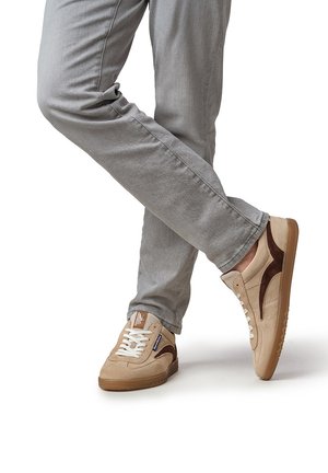 Person trägt hellgraue Hose und beigefarbene Turnschuhe mit braunen Akzenten, steht mit einem Bein über das andere geschlagen.