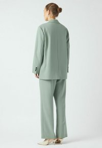 Grønn skreddersydd dress laget av glatt stoff; består av en lang blazer med notch-krage og bukser med vide ben. Spisse hvite hæler.