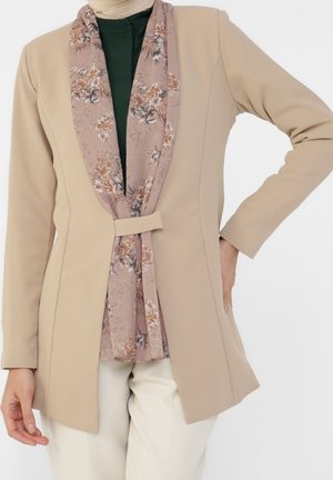 Frau trägt beige Blazer über dunkelgrünem Oberteil und einem floralen hellrosa Schal, mit cremefarbenen Hosen, posiert mit einer Hand in der Hüfte.