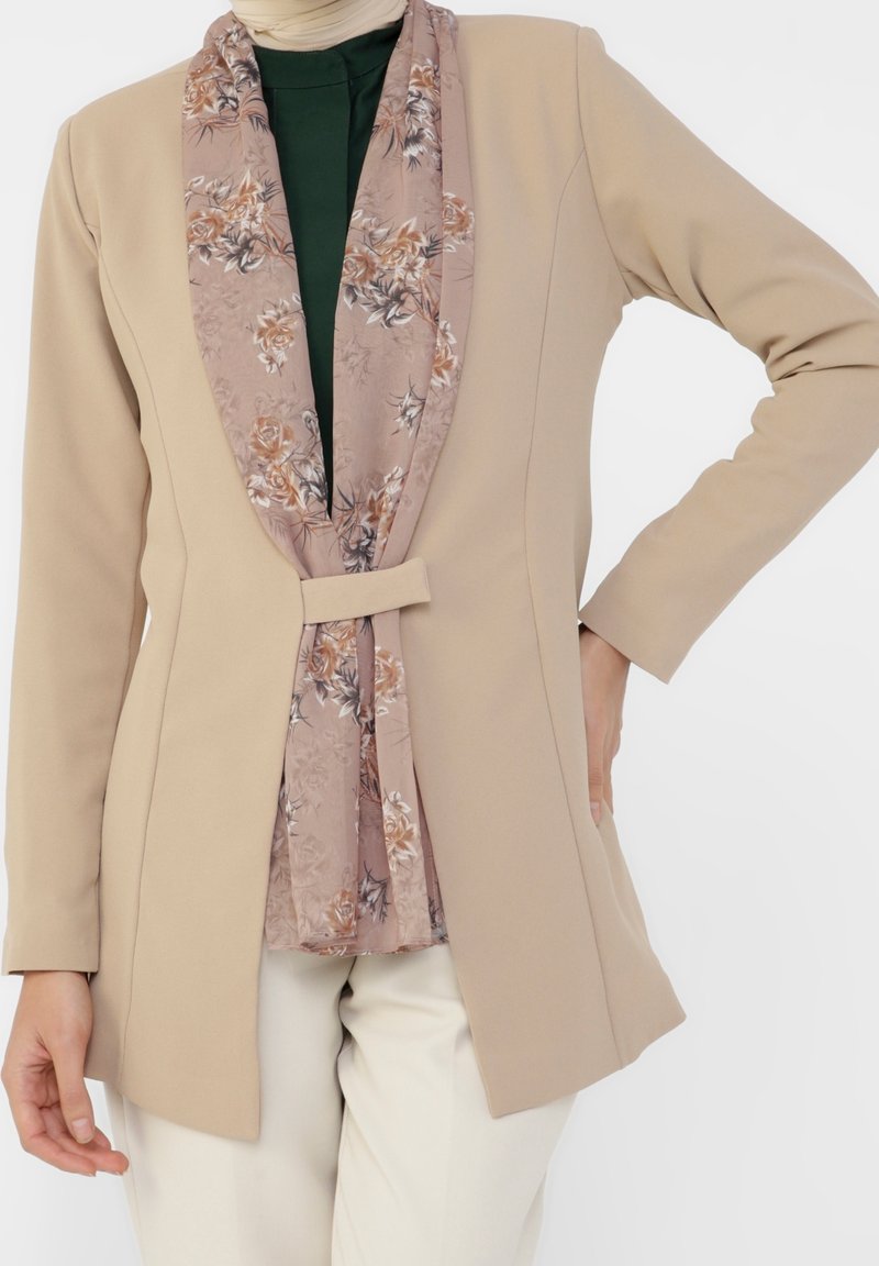 Femme portant un blazer beige sur un haut vert foncé et une écharpe fleurie rose clair, avec un pantalon de couleur crème, posant avec une main sur la hanche.