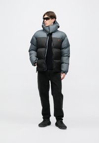 Pufová bunda v sivej a čiernej farbe s vysokým golierom, zapínaním na zips a nášivkou s logom na hrudi; v kombinácii s čiernymi džínsami a topánkami.