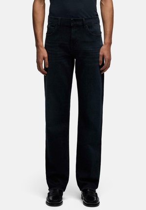 Personne portant un jean noir droit, une chemise noire et des chaussures noires, debout devant un fond uni.