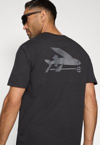 Čierne bavlnené tričko s grafickým dizajnom ryby s krídlami v sivej farbe na chrbte. Krátke rukávy a okrúhly výstrih.