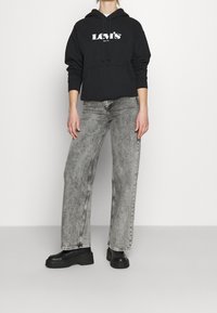 Sweat à capuche noir avec logo "Levi's" blanc, associé à un jean gris à jambes larges avec un motif délavé et des bottes plateformes noires.