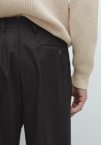 Mörkgrå byxor i texturerat tyg, med en bakficka med knappstängning, kombinerat med en ljusbeige ribbad tröja.