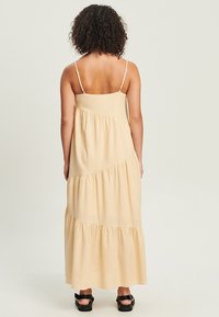 Maxi-mekko vaaleasta beige-kankaasta ohuilla olkaimilla, jossa on kerroksellisia röyhelöitä tekstuurin luomiseksi. Takana on sileä pinta suoran leikkauksen kanssa.