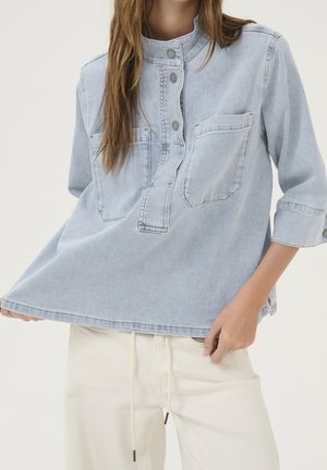 Kobieta ubrana w jasnoniebieską koszulę jeansową z zatrzaskami i dwoma kieszeniami na piersi, w połączeniu z kremowymi spodniami z troczkiem.