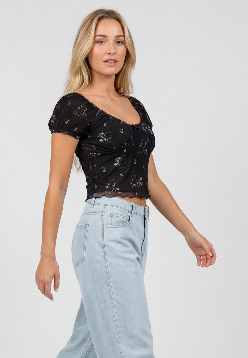 Femme marchant, portant un haut transparent à manches courtes avec un motif floral noir et un jean taille haute bleu clair, sur un fond gris uni.