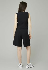 Femme se tenant dos à nous, portant un haut sans manches à rayures noires et un short au genou, avec des baskets blanches sur un fond gris.
