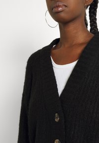 Woman wearing large hoop earrings, black textured cardigan with buttons, and white ribbed top, shown from neck to mid-torso.