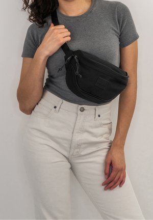 Femme portant un t-shirt gris ajusté à manches courtes et un pantalon clair taille haute avec un sac banane noir porté en bandoulière sur sa poitrine.