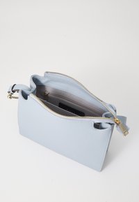 Borsa a mano in pelle azzurra chiara con superficie testurizzata, chiusura con zip e dettagli in metallo dorato. L'interno presenta una tasca e un logo.
