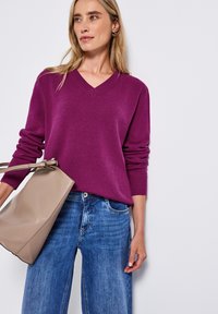 Pull violet en V avec des poignets côtelés, associé à un jean large bleu clair. Tient un sac fourre-tout beige en texture lisse.
