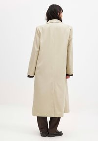 Beige long coat with a straight cut, large collar, and two side slits. Material appears smooth, paired with dark brown trousers and shoes.