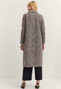 Langer Hahnentrittmantel mit Kragen, aus strukturiertem Stoff in Grau und Schwarz. Getragen über einer weit geschnittenen Hose und schwarzen Sandalen.