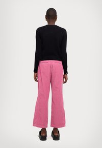 Maglione nero lavorato a maglia con maniche lunghe abbinato a pantaloni a gamba larga in velluto a coste rosa. Dettagli notevoli includono il tessuto testurizzato e le tasche posteriori.