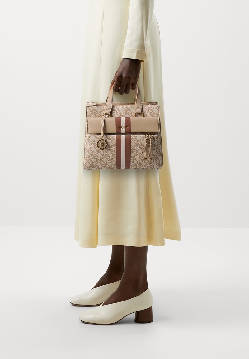 Beżowa torebka wzorzysta z dwoma uchwytami, jasnoróżowym paskiem, złotymi akcentami i detalem w postaci frędzli. Wykonana z miękkiej skóry i o sztywnej formie.