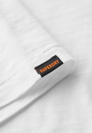 Wit textiel met een zachte, gestructureerde oppervlakte, voorzien van een zwart label met de opdruk "SUPERDRY" in oranje.