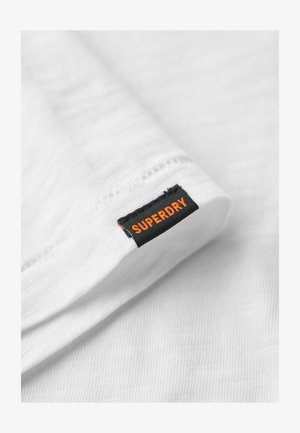 Wit textiel met een zachte, gestructureerde oppervlakte, voorzien van een zwart label met de opdruk "SUPERDRY" in oranje.