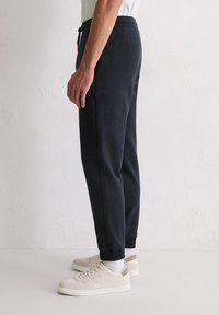 Dunkelgraue Jogginghosen mit elastischen Bündchen, ausgestattet mit einem Tunnelzugbund und feiner Struktur. Gekleidet mit beigen Sneakers und weißen Socken.