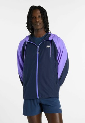 Mand med dreadlocks iført en marineblå og lilla New Balance lynlåstrøje og marineblå træningsshorts, stående mod en ensartet hvid baggrund.