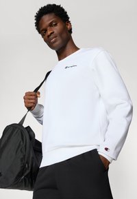 Sweatshirt blanc avec un col rond et un logo "Champion" noir brodé, associé à un pantalon noir et tenant un sac noir.