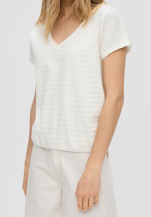 Kvinde iført en hvid, kortærmet tekstureret V-hals top og lyse bukser, stående mod en ensartet lys baggrund.