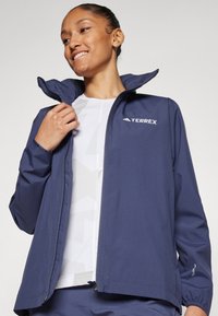 Jachetă din softshell de culoare albastru marin, cu guler înalt, închidere cu fermoar și logo. Prezintă manșete elastice, material ușor și design respirabil.