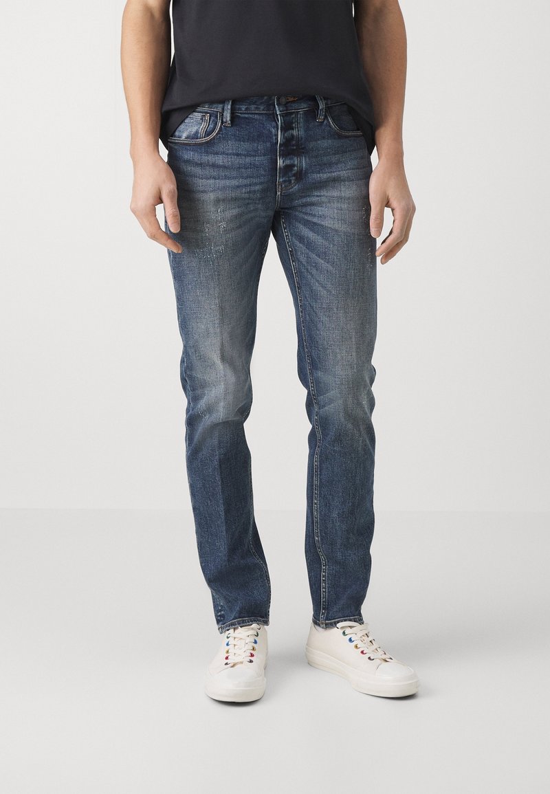Blå denimjeans med raka ben, med subtila blekningar och slitningar. Bärs med vita sneakers och en svart t-shirt.