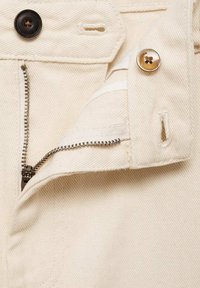 Tissu en denim beige avec une fermeture éclair métallique partiellement ouverte, deux boutons marron et des boutonnières visibles sur un vêtement.