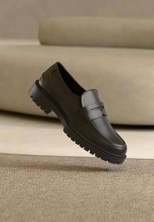 Mocassin en cuir noir avec semelle épaisse à crampons, détail de bride sur le dessus, posé sur un fond de tapis beige et canapé.
