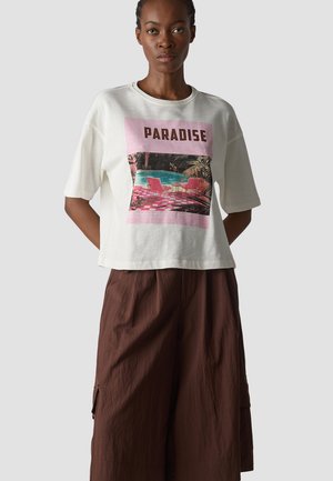 Woman wearing white t-shirt with pink "PARADISE" print and pool scene, paired with loose dark brown wide-leg pants, standing against gray background.