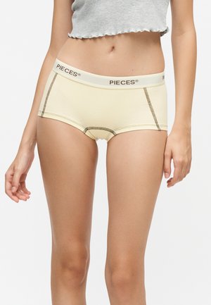 Kvinde iført beige hipster- undertøj med "PIECES" linning og synlige sømme, kombineret med en kort grå top.